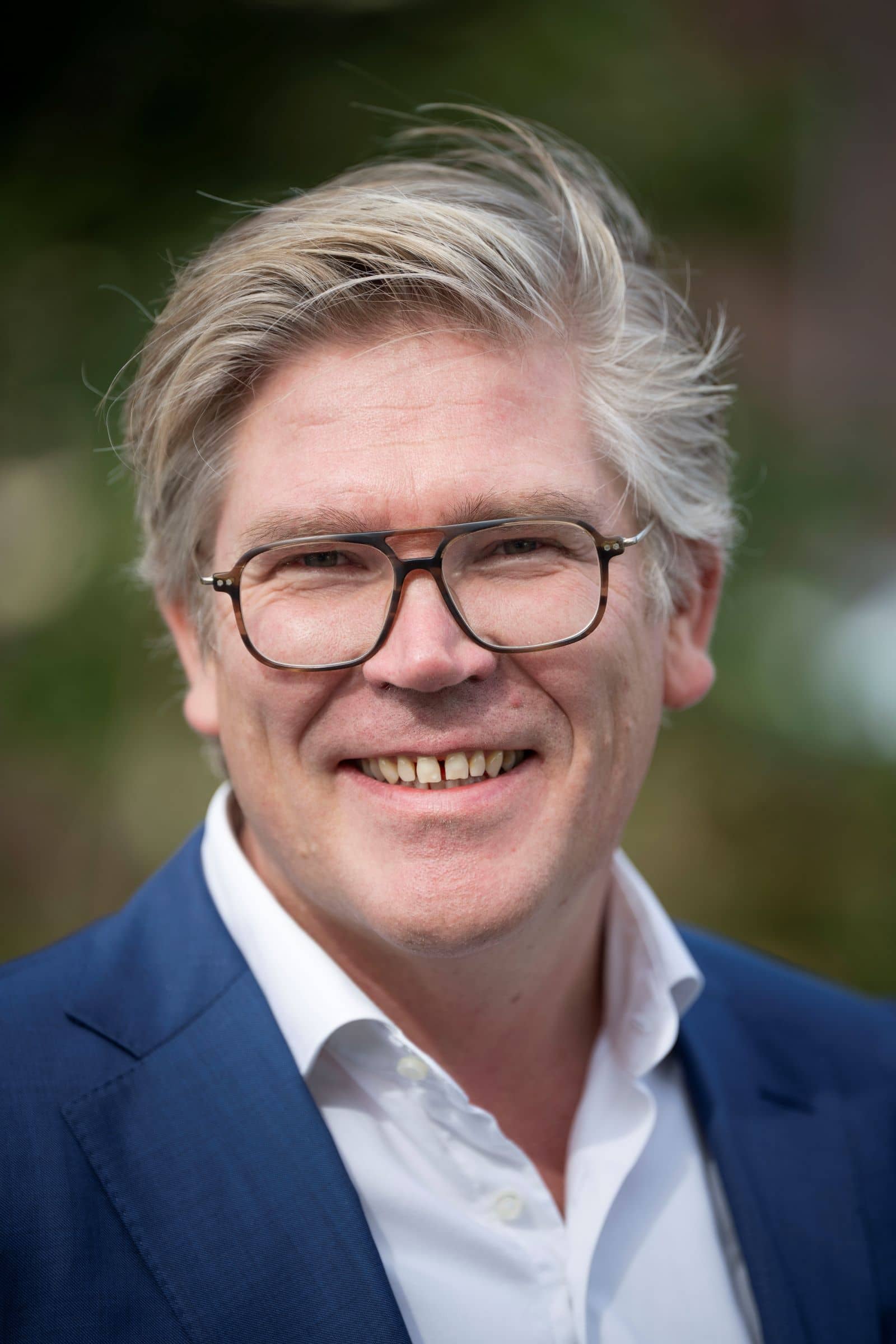 Een man met een donkere bril en grijs haar kijkt lachend naar de camera